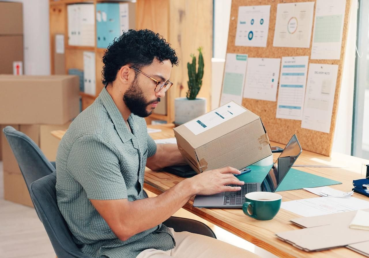 A man is holding a delivery box and typing on a laptop.
